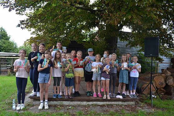 Zdjęcie grupowe dzieci na podwórku z gadżetami przekazanymi przez urząd gminy: dzieci dostały linijki, drewniane piórniki, małe składane puzzle oraz grę domino