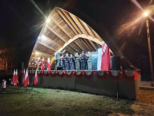 Scena koncertowa wieczorowa porą z zespołem muzycznym, w tle flaga biało czerwona oraz flagi pod sceną.