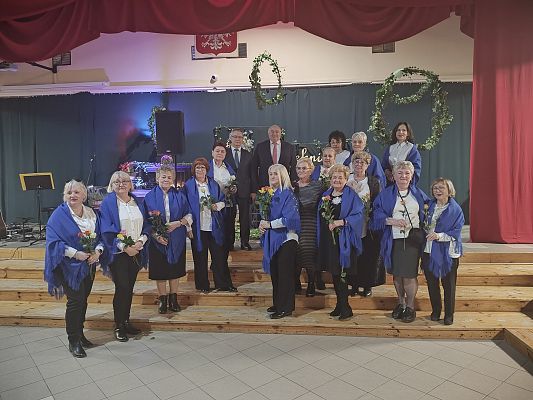 Panie z KGW Mosty na wspólnym zdjęciu z Wójtem Gminy Zdzisławem Chojnackim na tle zielonej dekoracji.