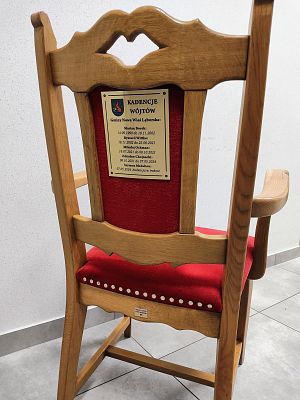 Zdjęcie przedstawia drewniane krzesło z tapicerowanym siedzisku a tle białej ściany